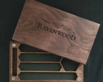 May include: A wooden box with a hinged lid, featuring the engraved text "Ravenwood" and a flock of birds. The box is open, revealing a green felt interior with several hexagonal compartments. The box is made of dark wood.