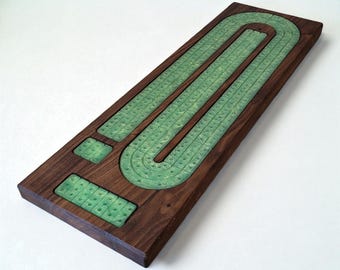 May include: A wooden cribbage board with a dark brown finish. The playing surface features a green, U-shaped track with numerous peg holes. Two smaller green areas are also present. The board is rectangular and designed for the card game cribbage.