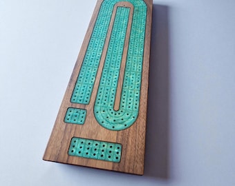 May include: Wooden cribbage board with a turquoise resin inlay. The board features three parallel tracks with numerous holes for pegging, and two scoring areas. The wood has a natural grain, and the resin has a marbled appearance.