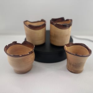 May include: Four handcrafted wooden cups with a natural, light brown finish. Each cup features a dark brown, organic-shaped rim. The cups are arranged on a black surface, showcasing their unique, rustic design.