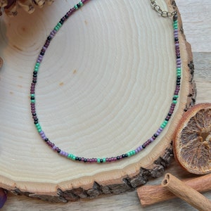 Könnte beinhalten: Eine zarte Choker-Kette aus kleinen Perlen in Violett-, Grün- und Schwarztönen. Die Kette liegt auf einer Holzoberfläche mit Tannenzapfen und Zimtstangen.