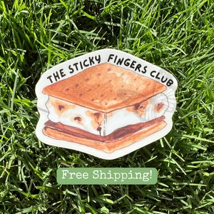 K&ouml;nnte beinhalten: Ein wei&szlig;er Aufkleber mit einer Cartoon-Illustration eines S'mores. Der S'more hat einen Graham-Cracker, eine ger&ouml;stete Marshmallow und Schokolade. Der Text "The Sticky Fingers Club" ist in Schwarz auf dem Aufkleber geschrieben.