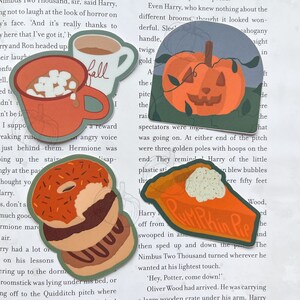 Może przedstawiać: Cztery kolorowe naklejki z motywami jesiennymi. Czerwony kubek z piankami i napisem "Fall", zielona dynia z twarzą jack-o'-lantern, stos pączk&oacute;w z pomarańczową i brązową polewą oraz kawałek ciasta dyniowego z białą polewą i napisem "Pumpkin Pie".
