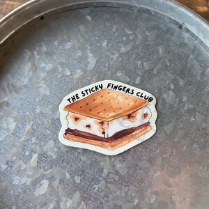 K&ouml;nnte beinhalten: Ein wei&szlig;er Aufkleber mit einer Cartoon-Illustration eines S'mores. Der S'more besteht aus einem Graham-Cracker, Schokolade und Marshmallow. Der Text "THE STICKY FINGERS CLUB" ist um den S'more herum geschrieben.