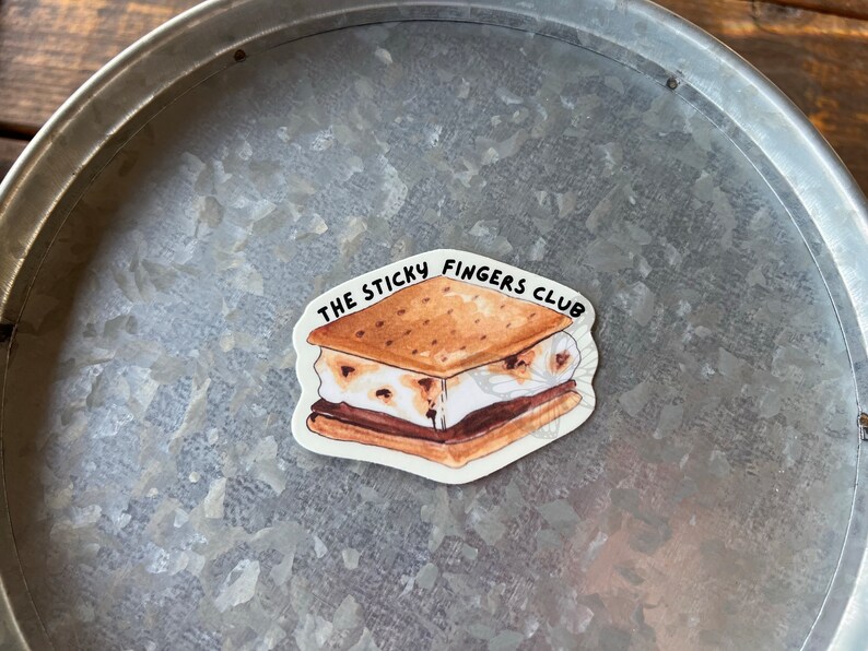 K&ouml;nnte beinhalten: Ein wei&szlig;er Aufkleber mit einer Cartoon-Illustration eines S'mores. Der S'more besteht aus einem Graham-Cracker, Schokolade und Marshmallow. Der Text "THE STICKY FINGERS CLUB" ist um den S'more herum geschrieben.