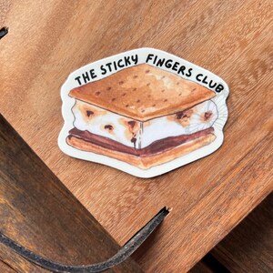 K&ouml;nnte beinhalten: Ein wei&szlig;er Aufkleber mit einer Cartoon-Illustration eines S'mores. Der S'more hat einen Graham-Cracker, Schokolade und Marshmallow. Der Text "THE STICKY FINGERS CLUB" ist in Schwarz um den S'more herum geschrieben.