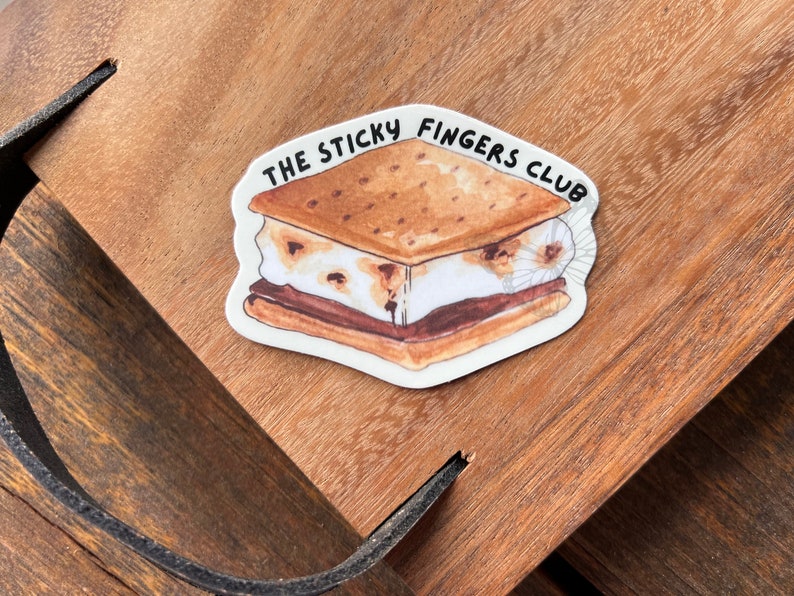 K&ouml;nnte beinhalten: Ein wei&szlig;er Aufkleber mit einer Cartoon-Illustration eines S'mores. Der S'more hat einen Graham-Cracker, Schokolade und Marshmallow. Der Text "THE STICKY FINGERS CLUB" ist in Schwarz um den S'more herum geschrieben.