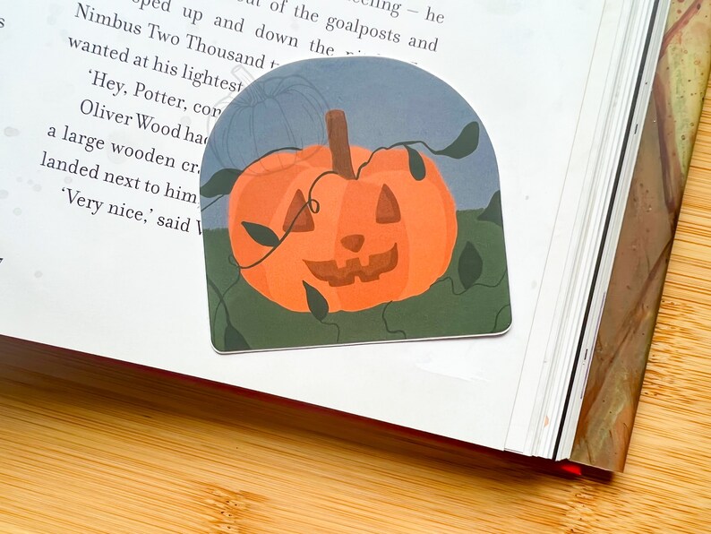 Może przedstawiać: Naklejka z motywem Halloween przedstawiająca wyrzeźbioną pomarańczową dynię z uśmiechającą się twarzą. Dynia jest otoczona zielonymi winoroślami i liśćmi, a tło jest jasnoniebieskie.