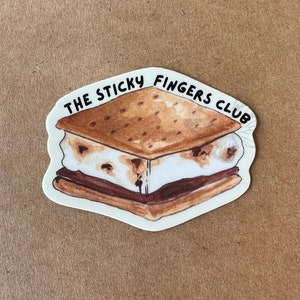 K&ouml;nnte beinhalten: Ein wei&szlig;er Aufkleber mit einer Cartoon-Illustration eines S'mores. Der Text "THE STICKY FINGERS CLUB" ist um den S'more herum geschrieben.