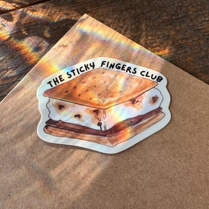 K&ouml;nnte beinhalten: Ein wei&szlig;er Aufkleber mit einer Aquarell-Illustration eines S'mores. Der S'more besteht aus einem Graham-Cracker, Marshmallow und Schokolade. Der Text "THE STICKY FINGERS CLUB" ist in Schwarz &uuml;ber dem S'more geschrieben.
