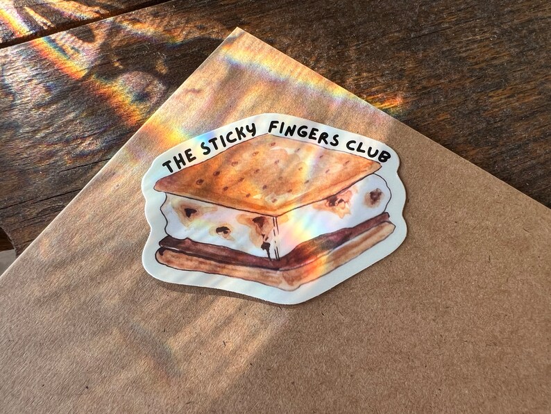 K&ouml;nnte beinhalten: Ein wei&szlig;er Aufkleber mit einer Aquarell-Illustration eines S'mores. Der S'more besteht aus einem Graham-Cracker, Marshmallow und Schokolade. Der Text "THE STICKY FINGERS CLUB" ist in Schwarz &uuml;ber dem S'more geschrieben.