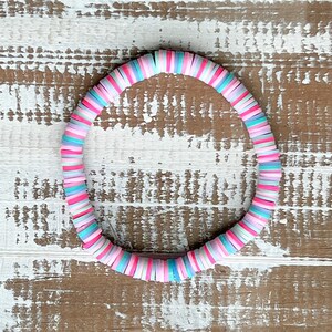 Könnte beinhalten: Ein farbenfrohes Armband aus runden, flachen Perlen in Rosa-, Blau- und Weißtönen. Das Armband ist auf einer Holzoberfläche kreisförmig angeordnet.
