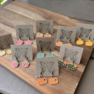 May include: A collection of pumpkin-shaped dangle earrings displayed on individual brown cardboard cards, each featuring a stamped butterfly logo. The earrings come in various colors including pink, orange, teal, and green, with some featuring polka dot patterns.