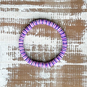 May include: A purple and white striped bracelet made of round beads. The bracelet is lying on a wooden surface.