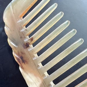 Puede incluir: Un peine para el cabello translúcido de color amarillo pálido con un acabado suave y pulido. El peine presenta un lomo curvo y sólido y una serie de dientes redondeados espaciados uniformemente. Se aprecian marcas naturales de color marrón y ámbar.