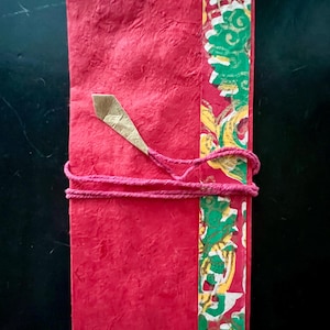 May include: A handmade red paper journal with a textured surface. The journal is bound with a pink cord and features a decorative strip with green, gold, and white patterns. A gold paper accent is attached to the cover.