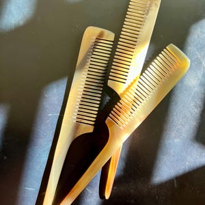 May include: Three hair combs crafted from natural horn material, featuring a translucent, marbled appearance with shades of cream, beige, and dark brown. Each comb has a long, tapered handle and fine, closely spaced teeth for grooming hair.