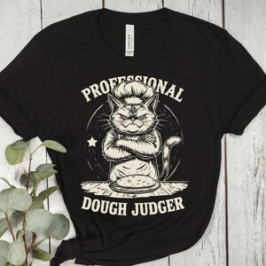 May include: Black t-shirt featuring a cartoon cat wearing a chef's hat with crossed arms. The text reads "PROFESSIONAL DOUGH JUDGER" in a circular design. The cat is positioned above a pizza. The shirt is made of a soft material.