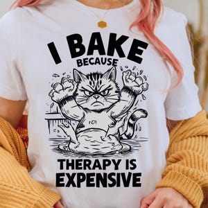 May include: White t-shirt with a black graphic of an angry cat in a baking scene. The text reads "I BAKE BECAUSE THERAPY IS EXPENSIVE". The cat is surrounded by flour and has an angry expression.