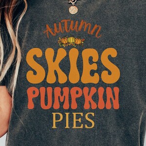 May include: A dark gray t-shirt with the text "Autumn Skies Pumpkin Pies" in a retro font. The text is in orange and yellow with a small graphic of pumpkins and leaves.
