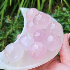 Puede incluir: Una colección de esferas de cuarzo rosa rosa en un plato blanco en forma de media luna. Las esferas son lisas y pulidas, con una apariencia translúcida.