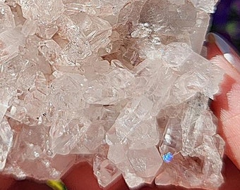May include: A cluster of clear quartz crystals with a pink hue. The crystals are arranged in a cluster, with some of the crystals pointing upwards. The crystals are translucent and have a slight sparkle.