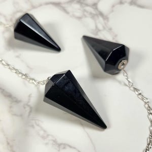 Puede incluir: Tres péndulos de cristal de obsidiana negra con cadenas plateadas. Los péndulos tienen la forma de una pirámide con una punta puntiaguda.