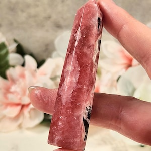 May include: A pink and black crystal point, also known as a wand or tower, is a polished piece of pink manganese calcite. The crystal is approximately 4 inches long and has a pointed tip.