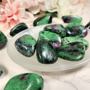 May include: A collection of green, black, and pink tumbled stones. The stones are smooth and polished, and they have a variety of shapes and sizes.