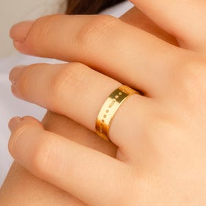 May include: A gold-colored ring with a series of small, evenly spaced dots engraved around the band. The ring is shown on a person's finger, with the hand and arm in the frame. The ring appears to be made of metal.
