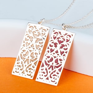 Op de afbeelding: Twee zilveren rechthoekige hangerkettingen met ingewikkelde opengewerkte ontwerpen. Eén hanger heeft een goudkleurige achterkant, de andere een roodkleurige achterkant. De kettingen worden gepresenteerd op een wit oppervlak tegen een oranje achtergrond.