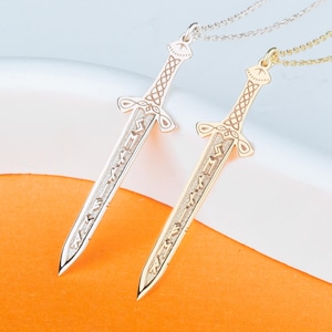 Puede incluir: Dos collares con colgantes de espada, uno plateado y otro dorado, se muestran sobre un fondo blanco y naranja. Los colgantes presentan intrincados diseños grabados y cuelgan de finas cadenas. Las espadas miden aproximadamente 5 cm de largo.
