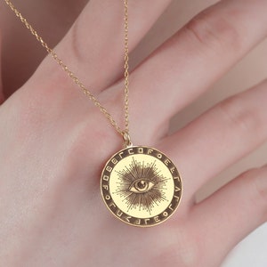 May include: A gold necklace with a round pendant featuring an eye with radiating lines and a circle of letters around the edge.