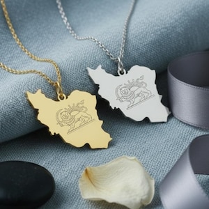 May include: Two necklaces with pendants shaped like the map of Iran. One is gold-colored, the other silver. Each pendant features a detailed emblem. The necklaces rest on a blue fabric with a gray ribbon, a black stone, and a white petal.