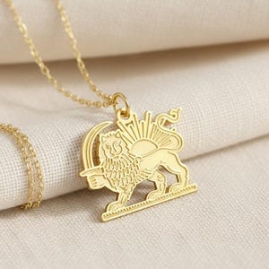 May include: A gold-colored pendant necklace featuring a lion with a sun emblem. The lion is standing on a rectangular base, with a sunburst design behind it. The necklace chain is delicate and gold-toned, resting on a cream-colored fabric.