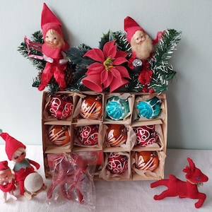 May include: Vintage Christmas ornament set featuring a box of glass ornaments in red, orange, and teal. The box is decorated with elf figurines, a poinsettia, and greenery. Additional red reindeer and elf figurines are also present.