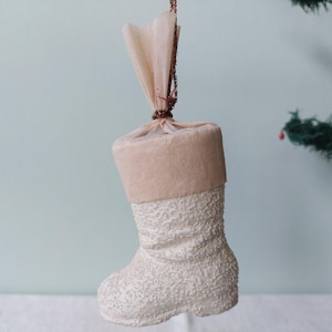 Op de afbeelding: Een vintage kerstsokornament. De sok is wit met een textuurpatroon en een beige manchet. Het hangt aan een bruin touwtje en is vastgebonden met een beige stof. De achtergrond is lichtblauw.