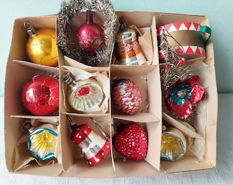 May include: A vintage box filled with assorted Christmas ornaments. The ornaments include gold, red, and silver baubles, a bell, a drum, and a flower. The box is divided into compartments, and the ornaments are nestled in tissue paper.