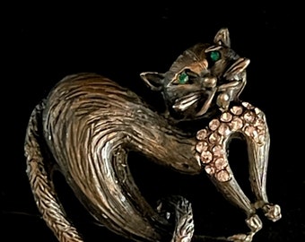 May include: A silver-toned cat brooch with green rhinestone eyes and a rhinestone collar. The cat is curled up with its tail wrapped around its body.