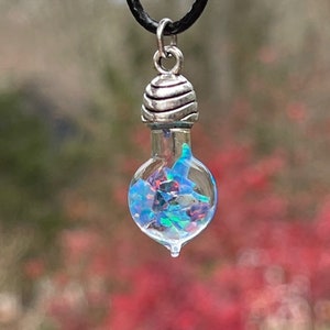 May include: A silver pendant necklace with a clear glass bottle filled with iridescent blue and green opal flakes. The bottle has a silver cap with a textured design. The necklace is on a black cord.