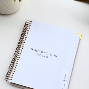 May include: A white spiral-bound "Daily Wellness Journal" with gold corner protectors. The journal has labeled tabs for "Journal", "Notes", and "Glossary". A small potted plant and a beige knitted item are in the background.