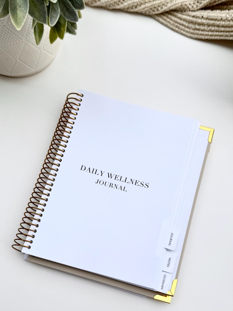 May include: A white spiral-bound "Daily Wellness Journal" with gold corner protectors. The journal has labeled tabs for "Journal", "Notes", and "Glossary". A small potted plant and a beige knitted item are in the background.