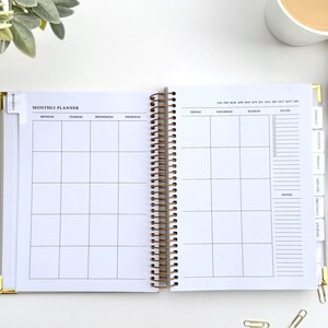 May include: Open monthly planner with a gold spiral binding. The planner is open to a blank monthly calendar layout with sections for each day of the week. Tabs on the side are labeled "Finance", "Notes", and more. A cup of coffee and a beige sweater are in the background.