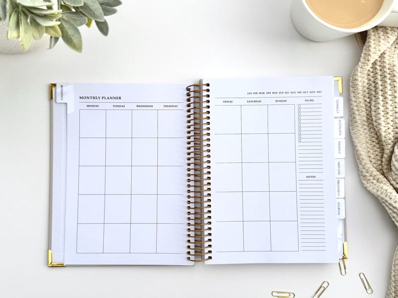 May include: Open monthly planner with a gold spiral binding. The planner is open to a blank monthly calendar layout with sections for each day of the week. Tabs on the side are labeled "Finance", "Notes", and more. A cup of coffee and a beige sweater are in the background.