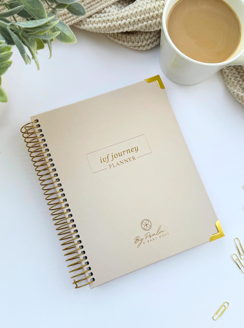 May include: A beige spiral-bound planner with gold accents, labeled "ivf journey PLANNER." The planner features gold corner protectors and a gold spiral binding. The cover includes the text "By Paola Baby Dust" and a floral design.