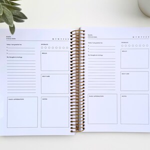 May include: Open spiral-bound planner with gold accents. The pages are white with black text and include sections for daily affirmations, notes, and tracking hydration and meals. The planner is open to a double-page spread.