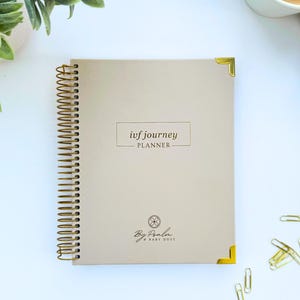 May include: A beige spiral-bound planner with gold accents, featuring the text "ivf journey PLANNER." The planner has gold corner protectors and a gold spiral binding. The bottom of the planner has the text "By Paola & BABY DUST."