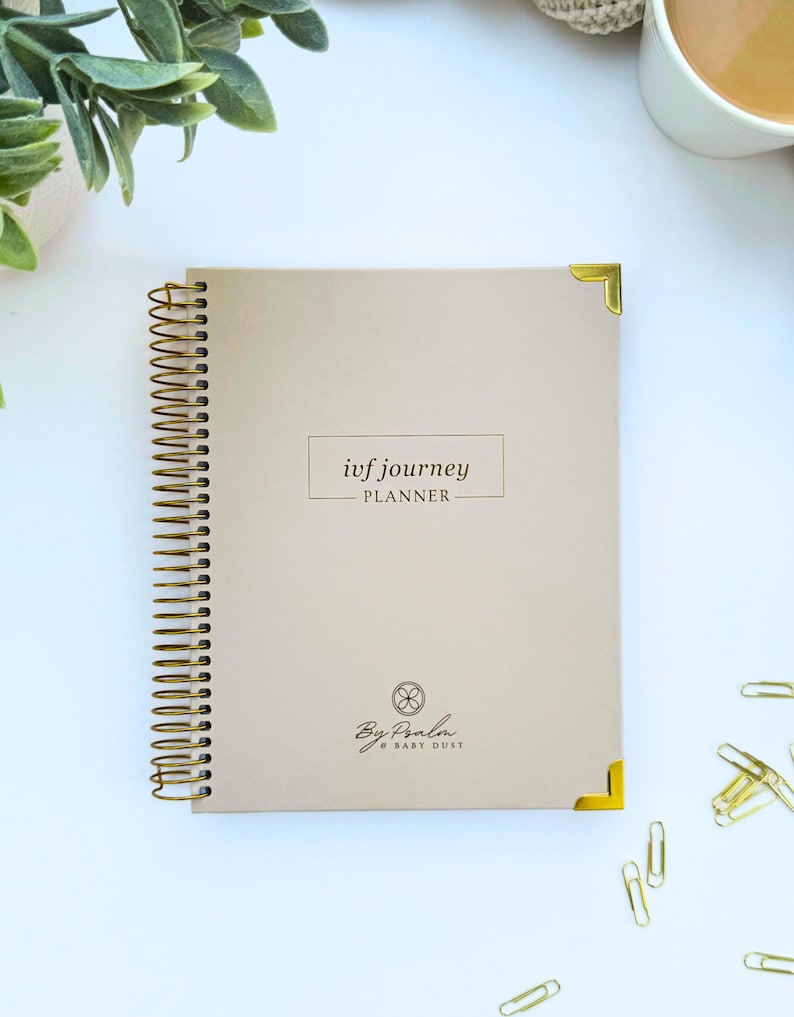 May include: A beige spiral-bound planner with gold accents, featuring the text "ivf journey PLANNER." The planner has gold corner protectors and a gold spiral binding. The bottom of the planner has the text "By Paola & BABY DUST."