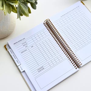 May include: Open white spiral-bound planner with gold accents, featuring a medication log. The planner is open to a page with a grid for tracking medication, dates, and times. Tabs for finance, accounts, and calendar are visible.