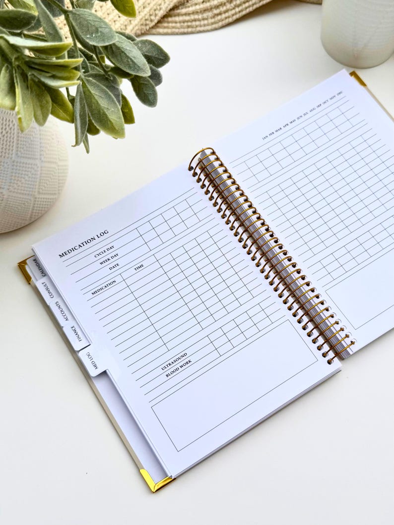 May include: Open white spiral-bound planner with gold accents, featuring a medication log. The planner is open to a page with a grid for tracking medication, dates, and times. Tabs for finance, accounts, and calendar are visible.